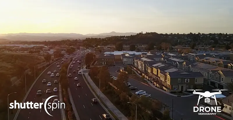 Drone aerial view of a subdivision at sunset, showing nearby multi-lane streets and mountain backdrop, captured by ShutterSpin. Aerial photography highlights both property context and neighborhood accessibility for real estate marketing.